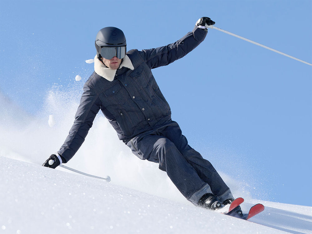 Men's model in action on the slopes wearing the Jeen ski jacket and Jett ski trousers in denim with a contrasting fur collar and selvedge details, paired with skis and ski poles.
