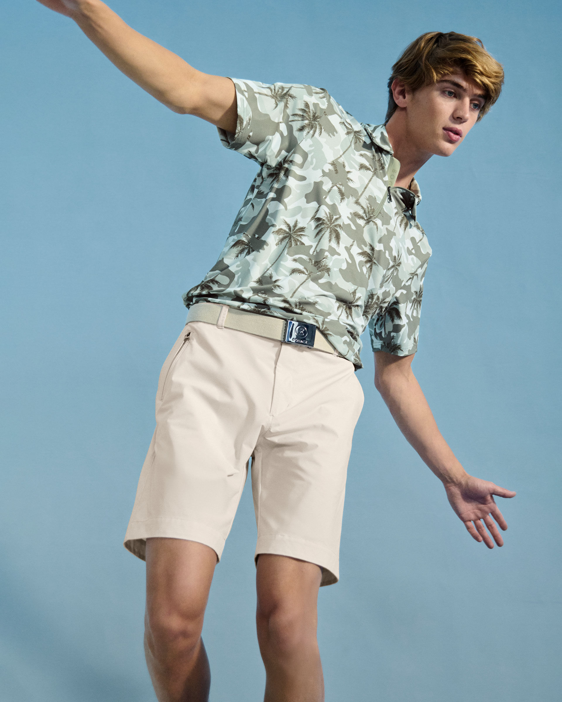 Das blonde Herren Model in dynamischer Pose tr&auml;gt ein Polo-Shirt mit Palmen- und Camouflage-Print, dazu eine Shorts und einen G&uuml;rtel.