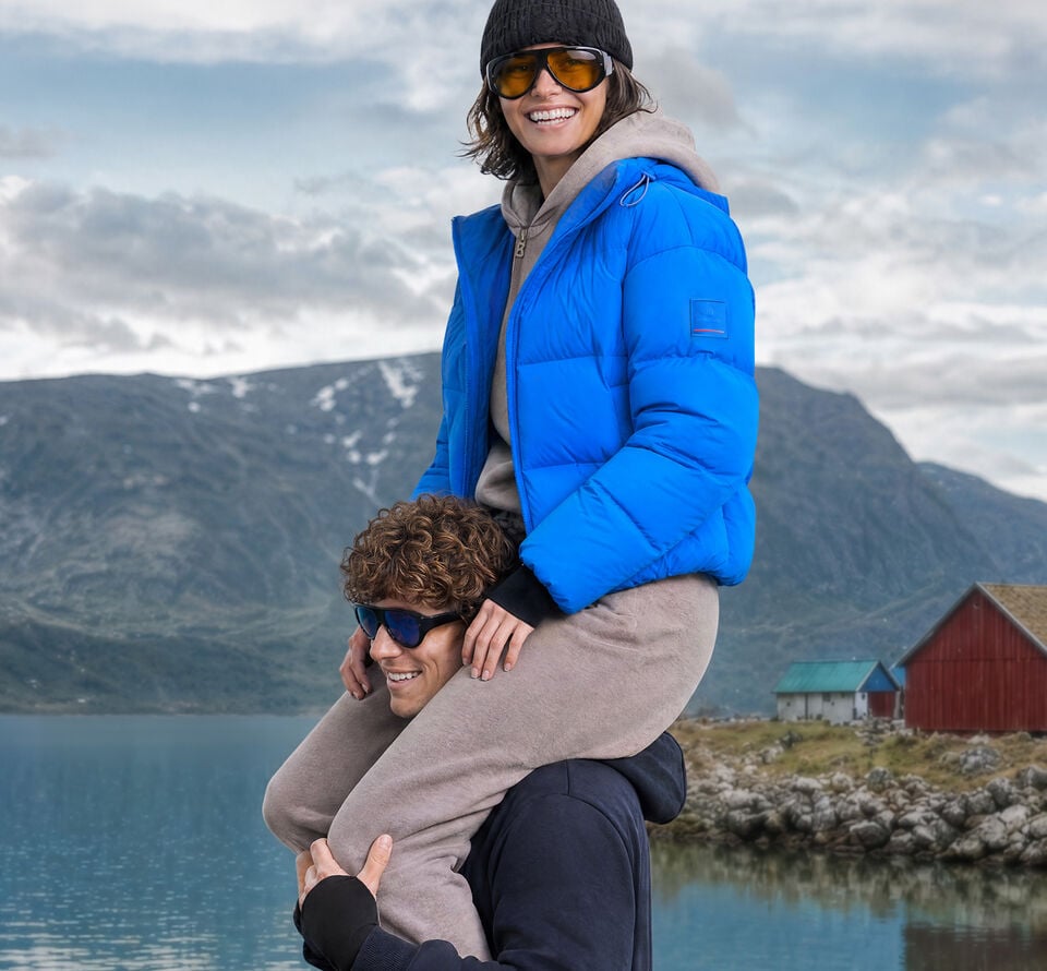 Bergkulisse. Das Herren Model tr&auml;gt das Damen Model auf den Schultern. Sie tr&auml;gt eine royalblaue Steppjacke, eine M&uuml;tze und eine Sonnenbrille.
