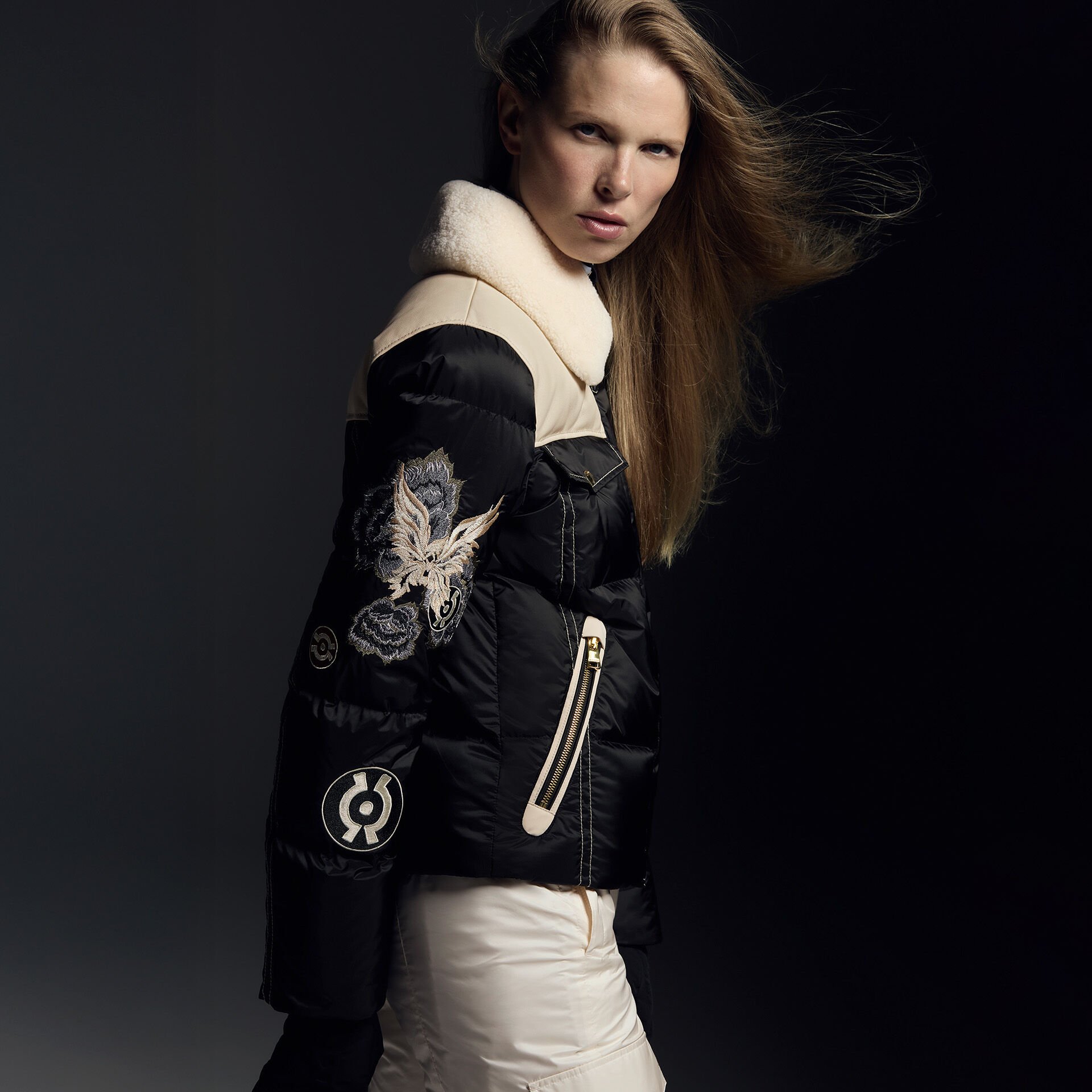 Das weibliche Model trägt eine beige glänzende Skihose und eine schwarze Skijacke mit Adler-Motiv und beigem Shearling-Kragen.