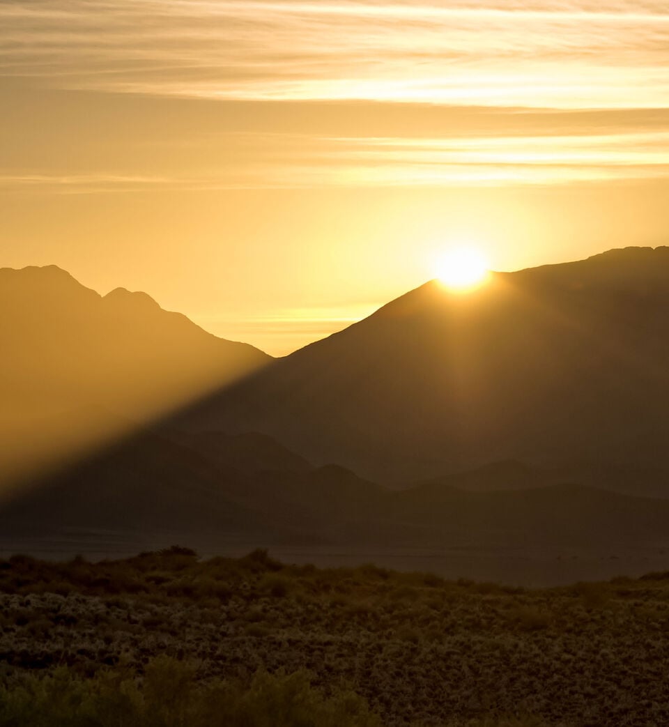 Idyllischer Sonnenuntergang hinter einer Berglandschaft vor einem Himmel in warmen Gelb- und Oranget&ouml;nen.
