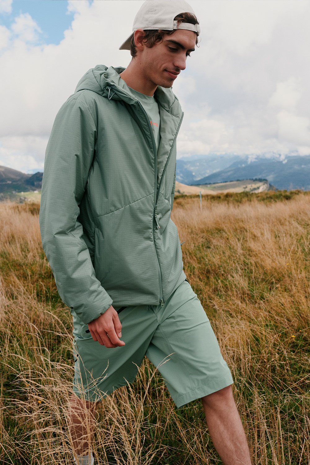 The male model is running through a grassy landscape with mountains in the background. He is wearing a total look in eucalyptus green.