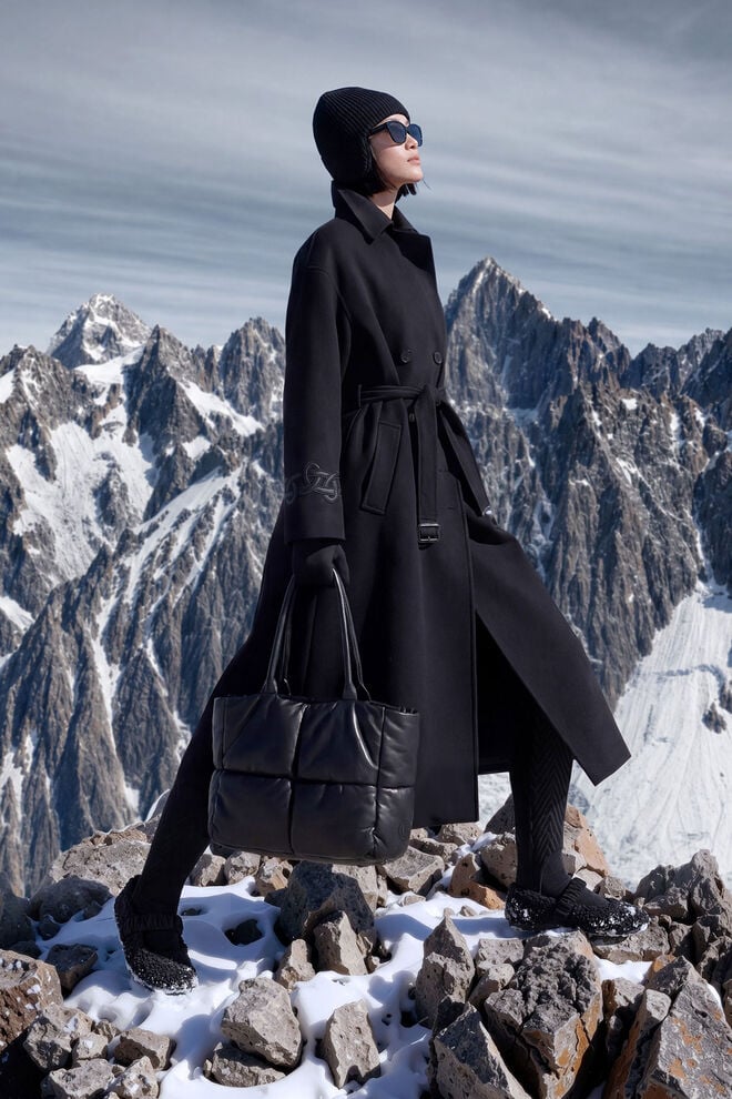 The female model with mountain scenery in the background is wearing a black coat with a wrap belt, and a knitted hat, sunglasses, a tote bag and teddy-fur ballerina flats to complete the look.