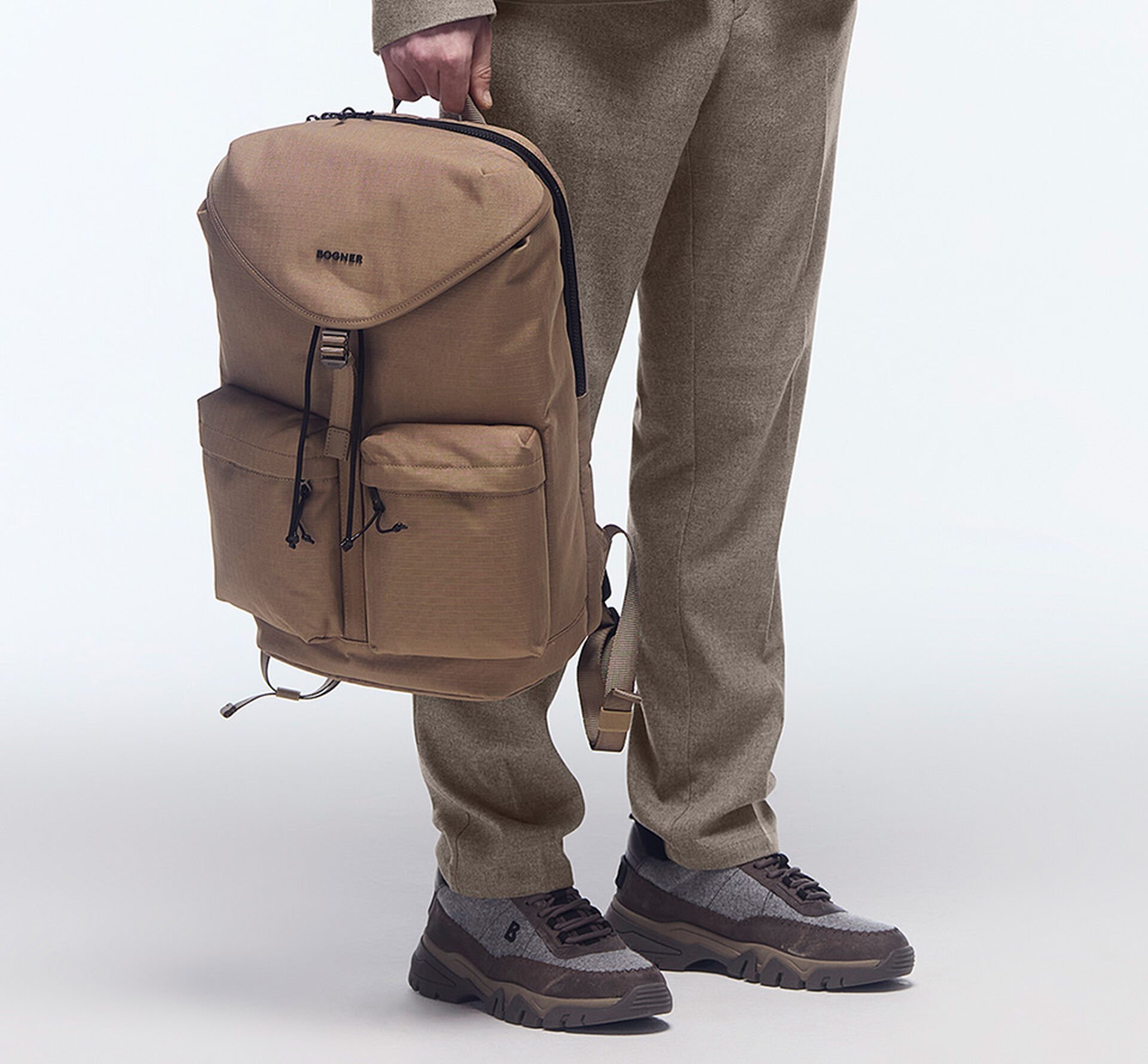 The male model in beige mottled trousers wears BOGNER sneakers and carries a spacious backpack in his hand.