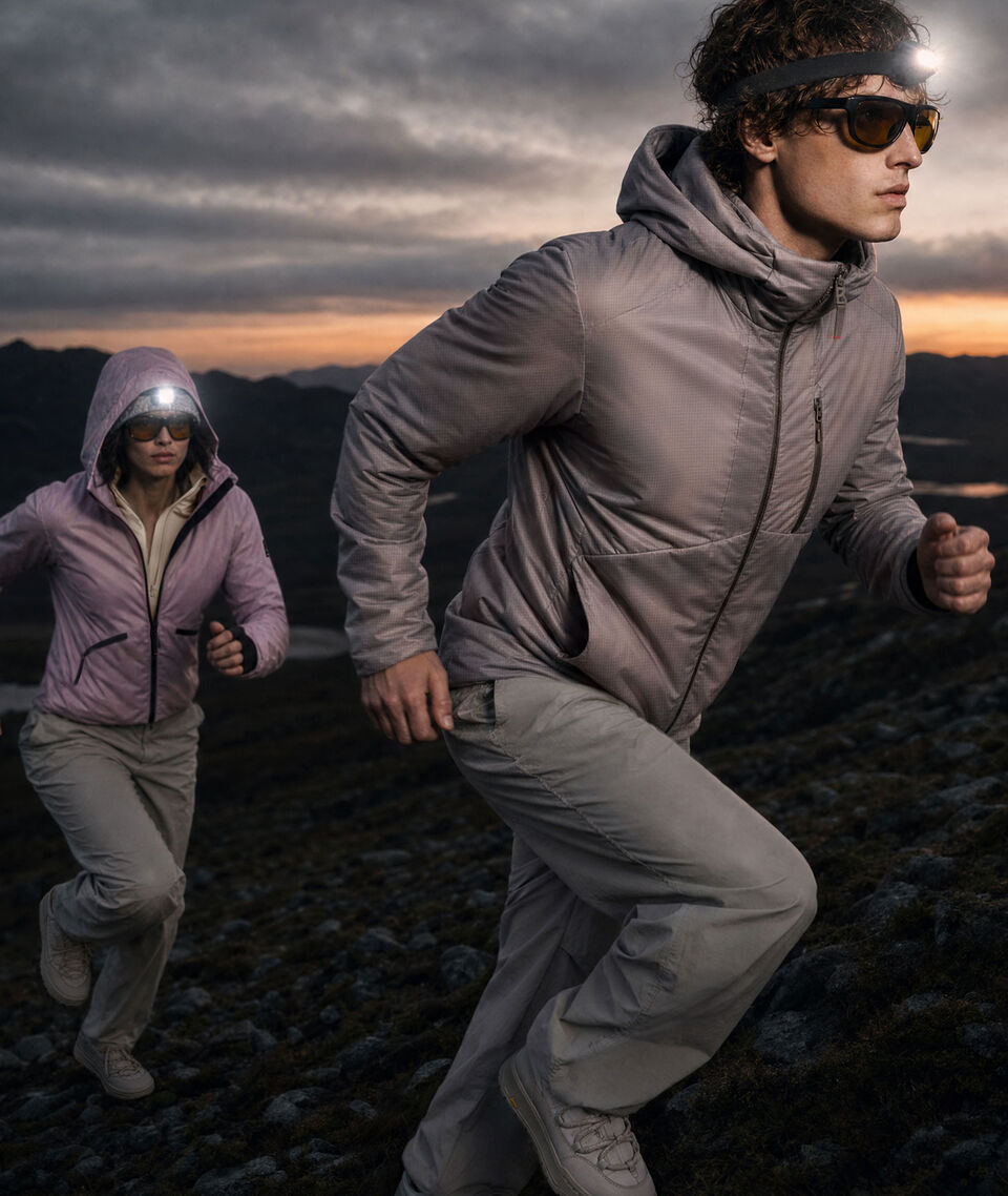 N&auml;chtliche Stimmung. Zwei vor einer Berglandschaft laufende Models, die FIRE+ICE Jacken und Hosen tragen. Von ihren K&ouml;pfen leuchten Stirnlampen.