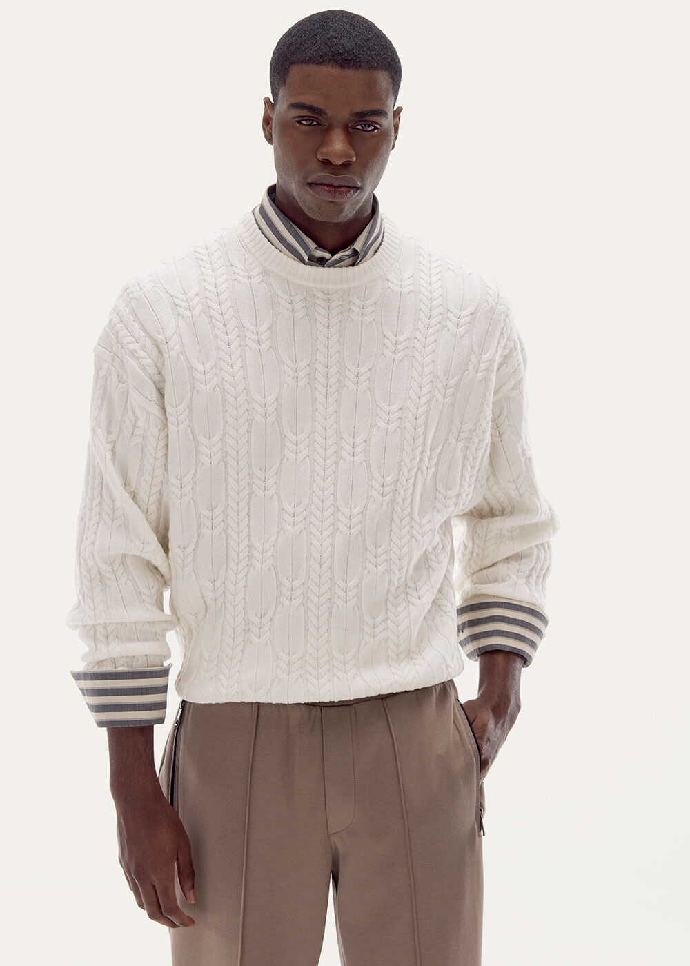 Halbkörperaufnahme mit weißem Zopfstrick-Pullover, darunter ein gestreiftes Hemd. Sweatpants komplettieren den Look.