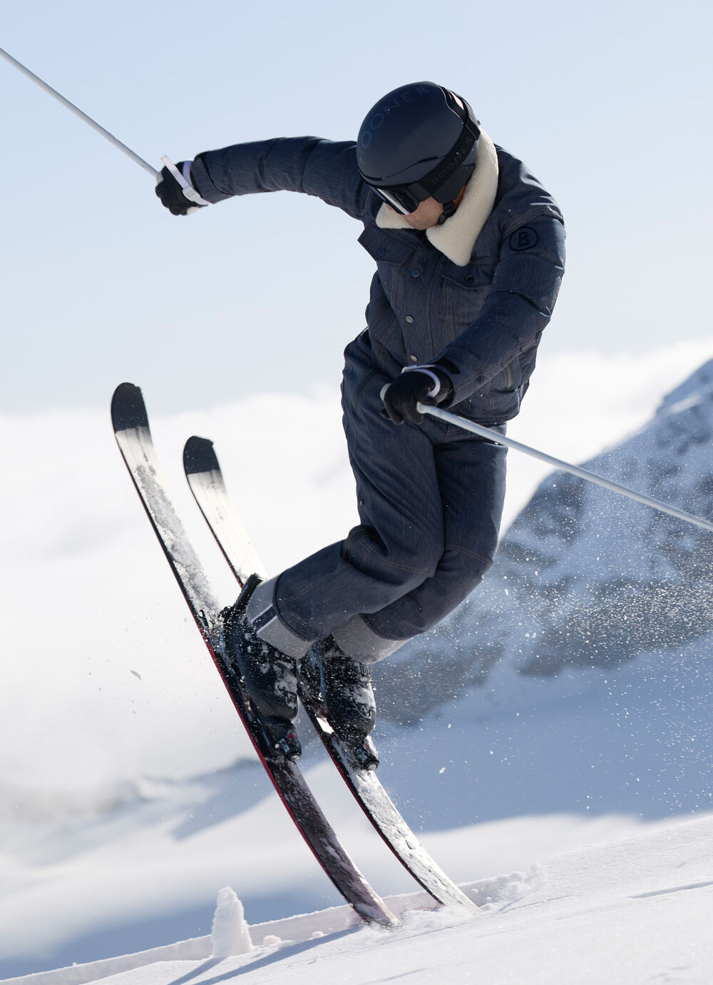 Men's model in action on the slopes wearing the Jeen ski jacket and Jett ski trousers in denim with a contrasting fur collar and selvedge details, paired with skis and ski poles.