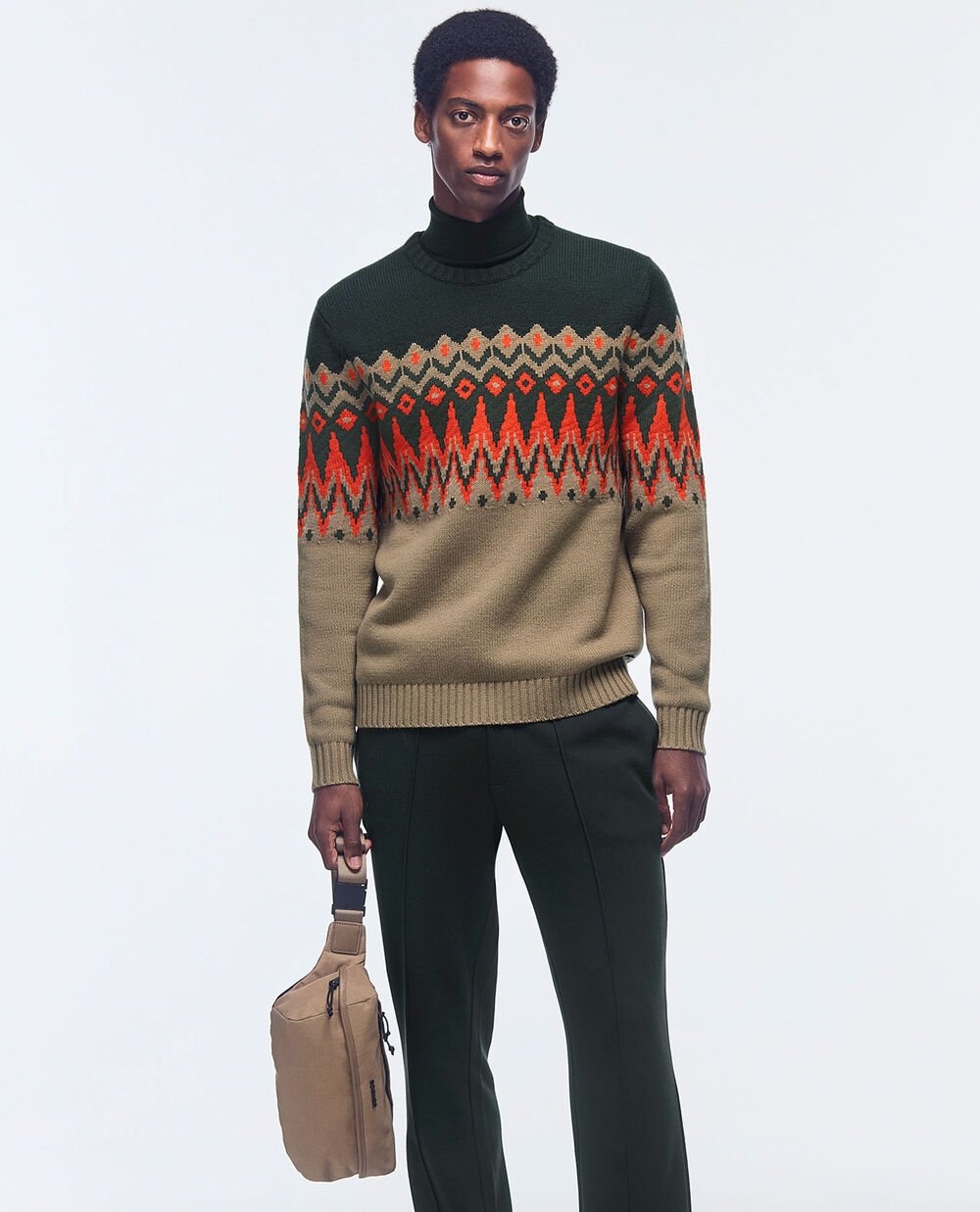 The model is wearing a dark green Norwegian sweater with beige and coral-coloured accents. Smart grey trousers complete the look.