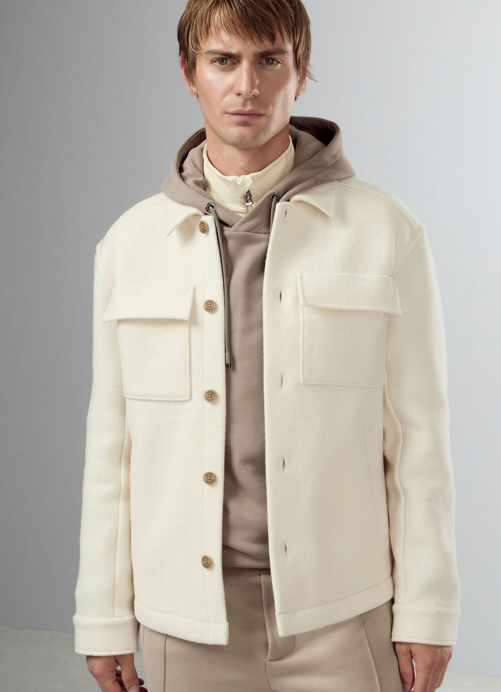 Half-body shot of the male model wearing a beige hoodie over a Troyer, topped with a shirt jacket.