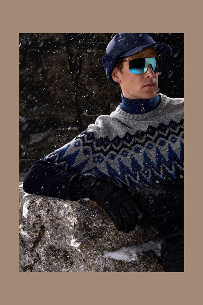The male model is sitting on snow-covered ground and wearing a Norwegian jumper with a blue, black and grey pattern.