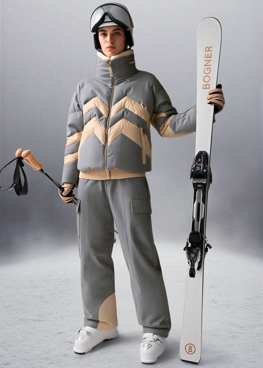 The female model is wearing an eucalyptus-colored ski outfit, a black ski helmet, and is holding skis with a B print.