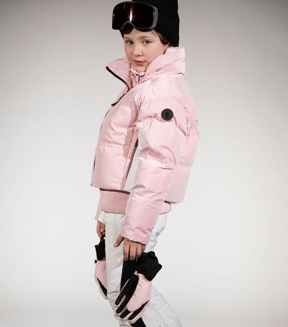 The girl is wearing a light pink ski jacket and holding matching gloves in her hand, paired with white ski trousers, a black beanie and ski goggles.
