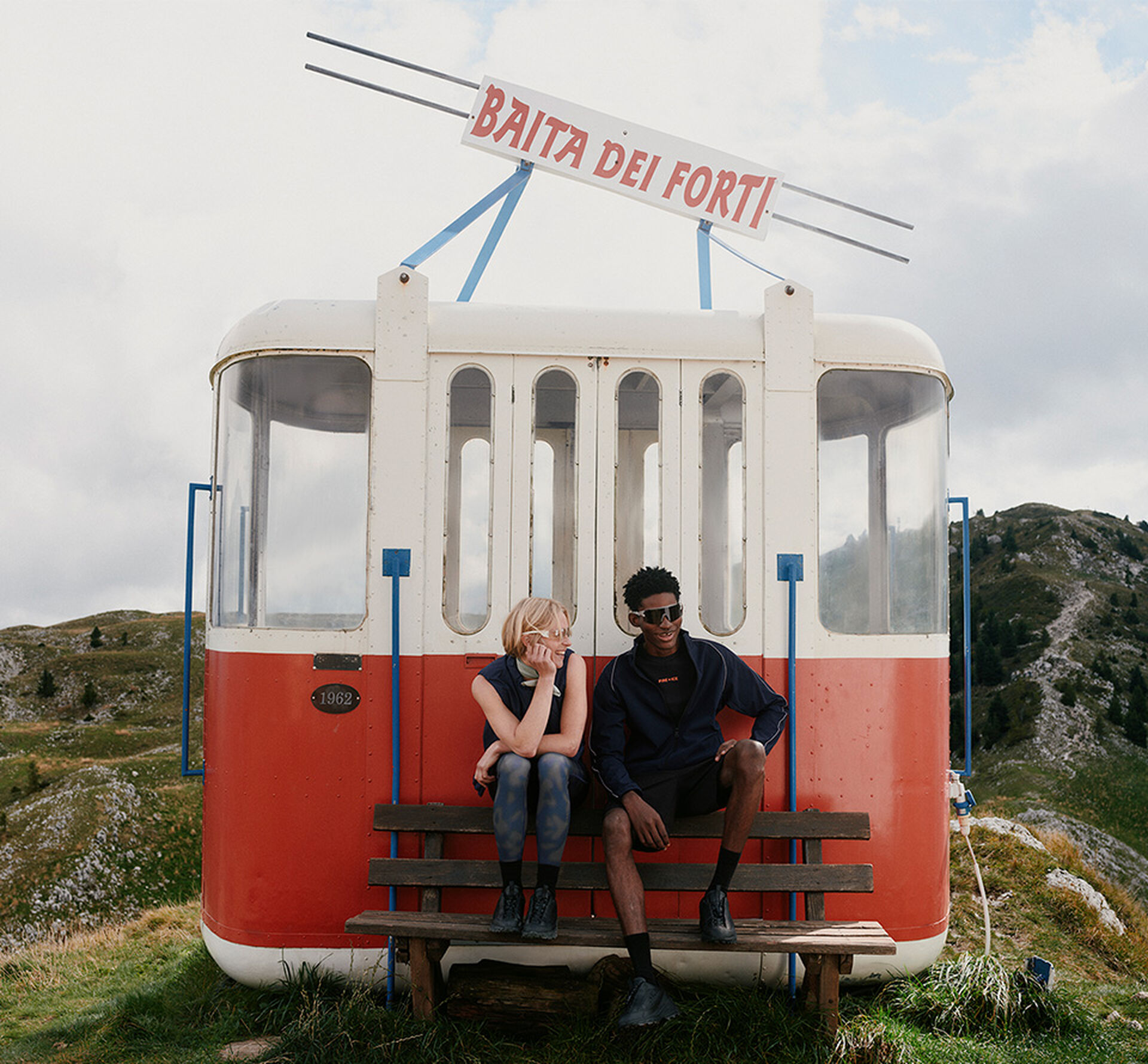 Aufnahme einer roten Skigondel am Berg, davor ein Damen und ein Herren Model in Sportswear auf einer Bank sitzend.