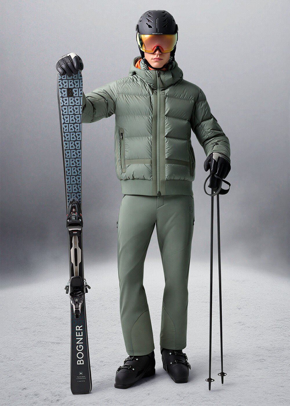 Das Damen-Model trägt ein eukalyptusfarbenes Ski-Outfit, einen schwarzen Skihelm und hält Ski mit B-Print in den Händen.