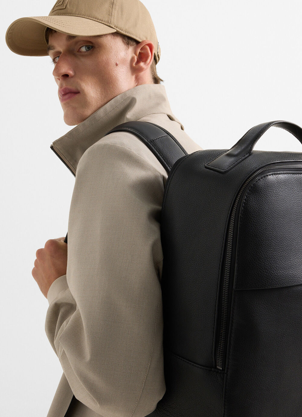The male model is seen from the back. He carries a beige baseball cap and a black backpack made from leather.