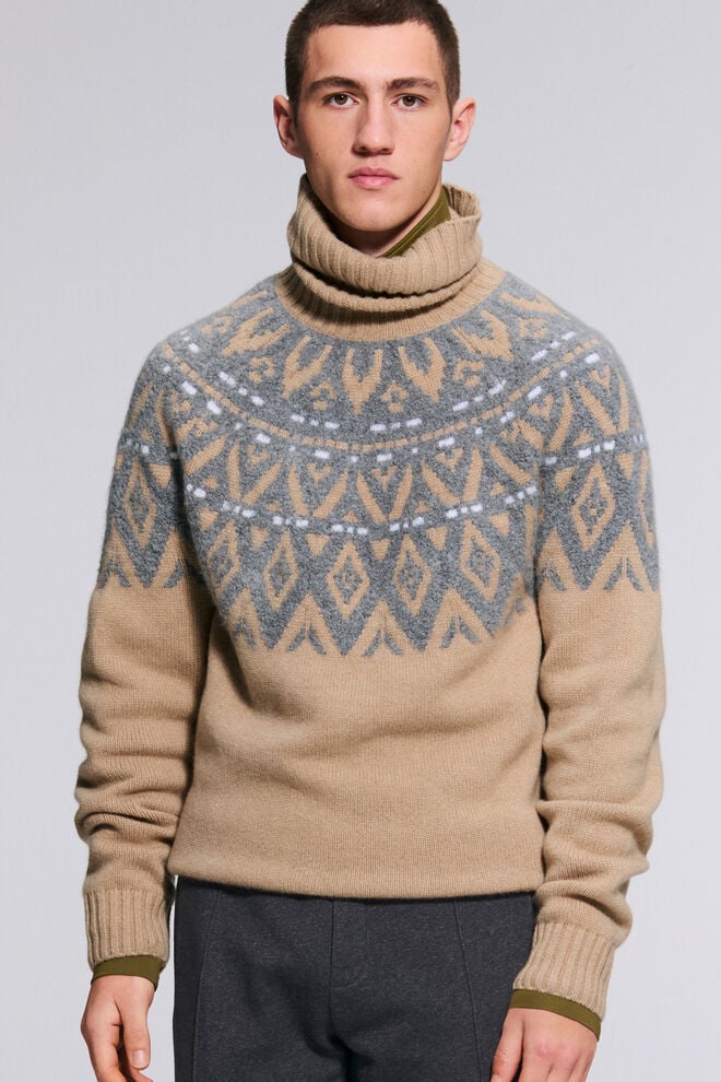 The model is wearing the beige Roxter pullover, which is embroidered with a grey and white Norwegian pattern.