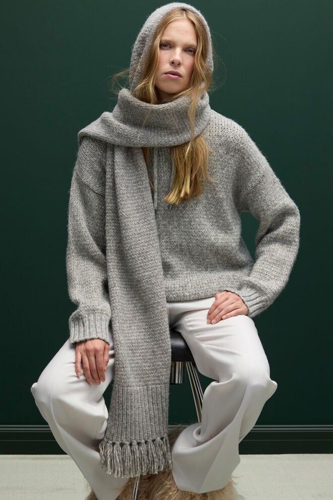 Le mannequin assis porte le sweat à capuche en maille Stella gris, assorti d'une écharpe de couleur coordonnée et d'un pantalon clair.