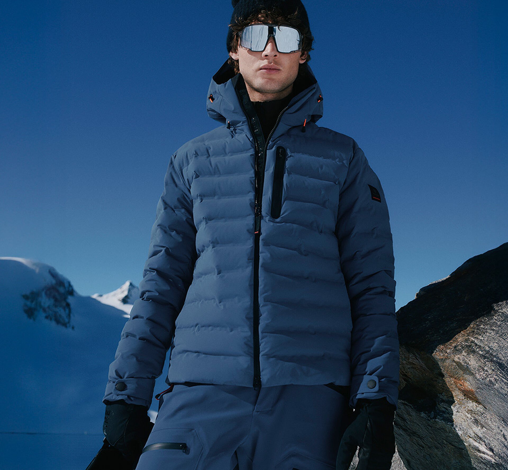 Das Herren Model trägt eine royalblaue Daunenjacke mit Steppung, schwarzen Logo-Details und Kapuze.