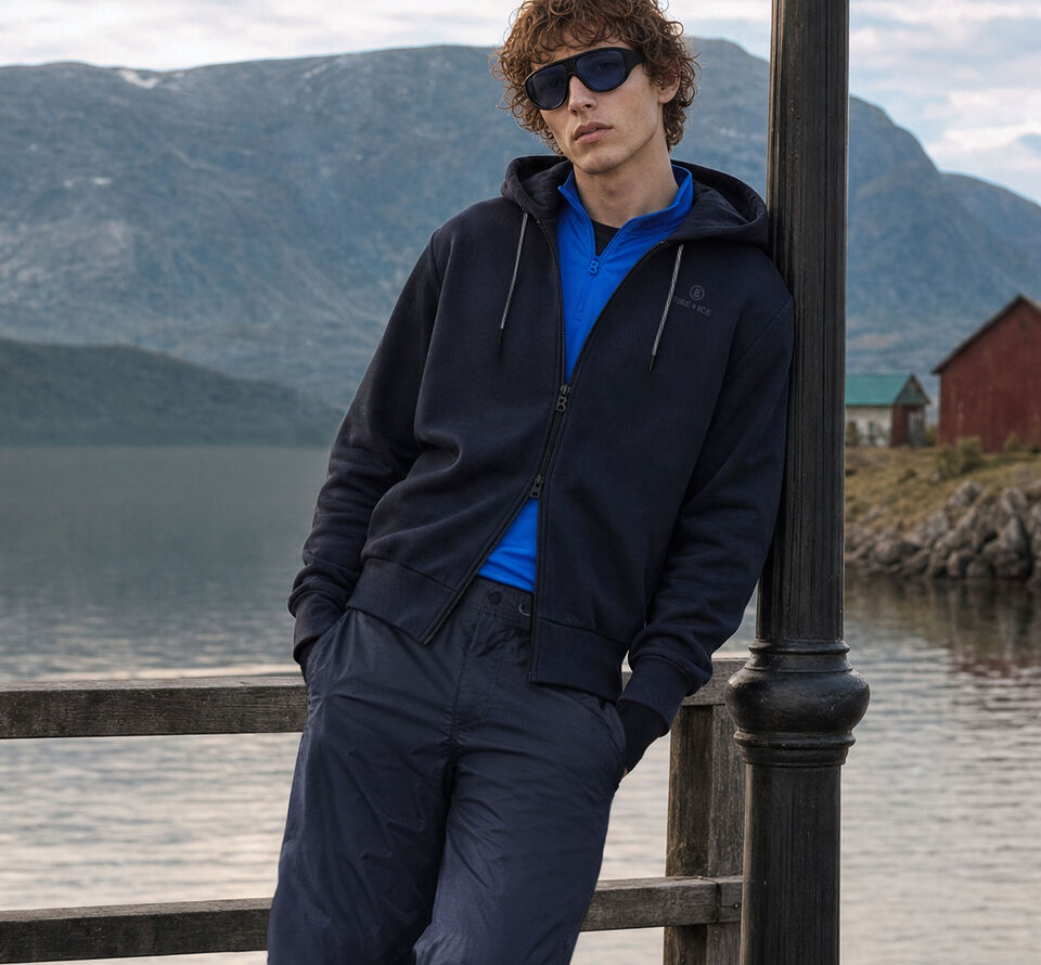 Das Herren Model steht vor einem Bergsee mit Gebirgskulisse. Er tr&auml;gt eine dunkelblaue Funktionsjacke und eine passende Hose, dazu First Layer Pascal darunter.