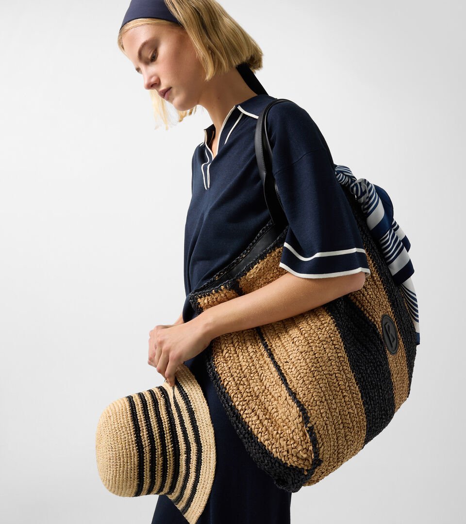 Seitliche Aufnahme des Damen Models. Sie tr&auml;gt ein navy-blaues Set sowie eine Tasche und einen Hut aus Bast in den Armen.