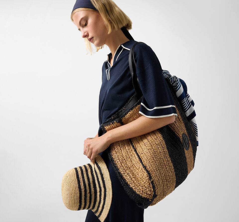 Seitliche Aufnahme des Damen Models. Sie tr&auml;gt ein navy-blaues Set sowie eine Tasche und einen Hut aus Bast in den Armen.