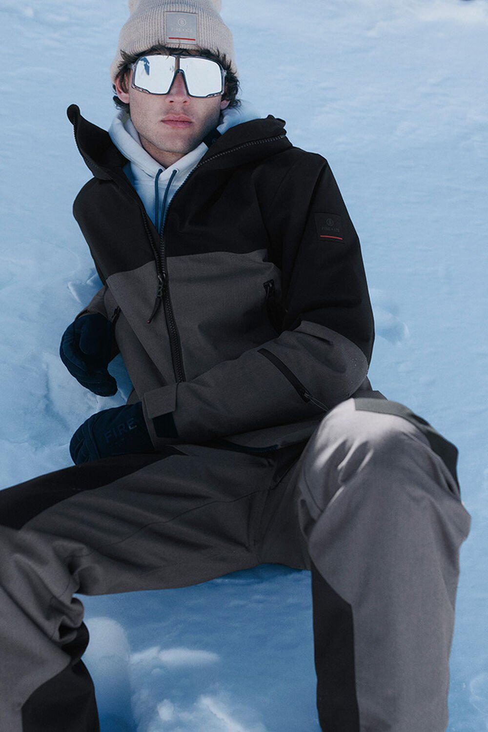 Das Herren Model lehnt im Schnee und trägt einen braun-schwarzen Ski Look mit Hoodie, Mütze und Skibrille.
