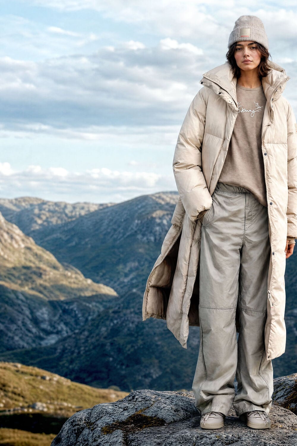Das Damen Model in einer Berglandschaft tr&auml;gt Daunenmantel Fleury, darunter T-Shirt Kate mit Ski-Print und Hose Paris aus technischen Materialien.