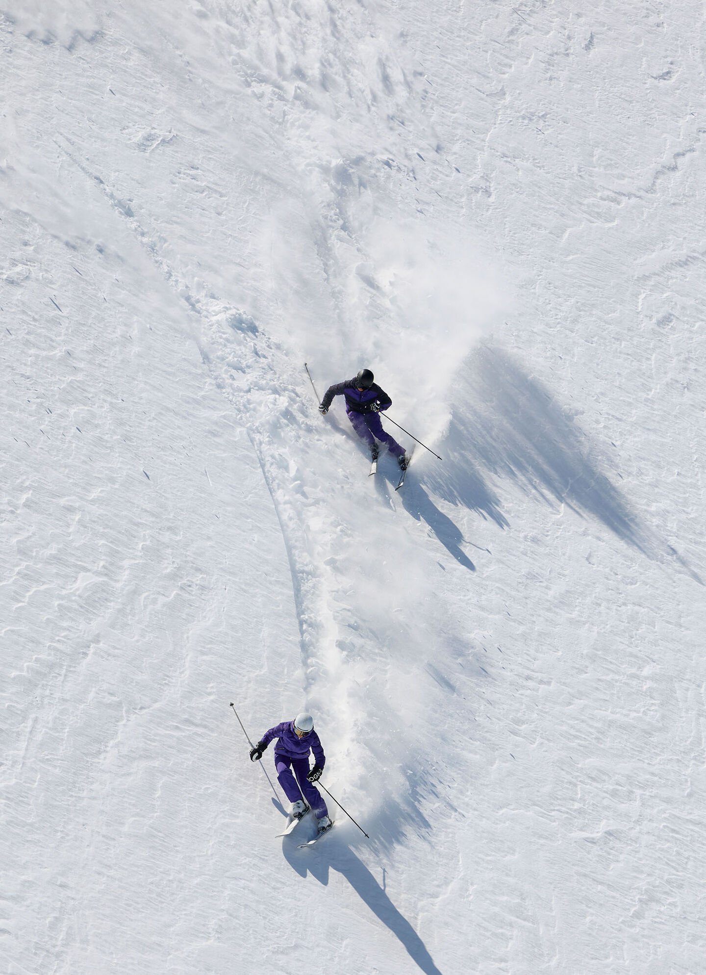 Zwei Skifahrer fahren die Piste runter und hinterlassen Spuren im Schnee.