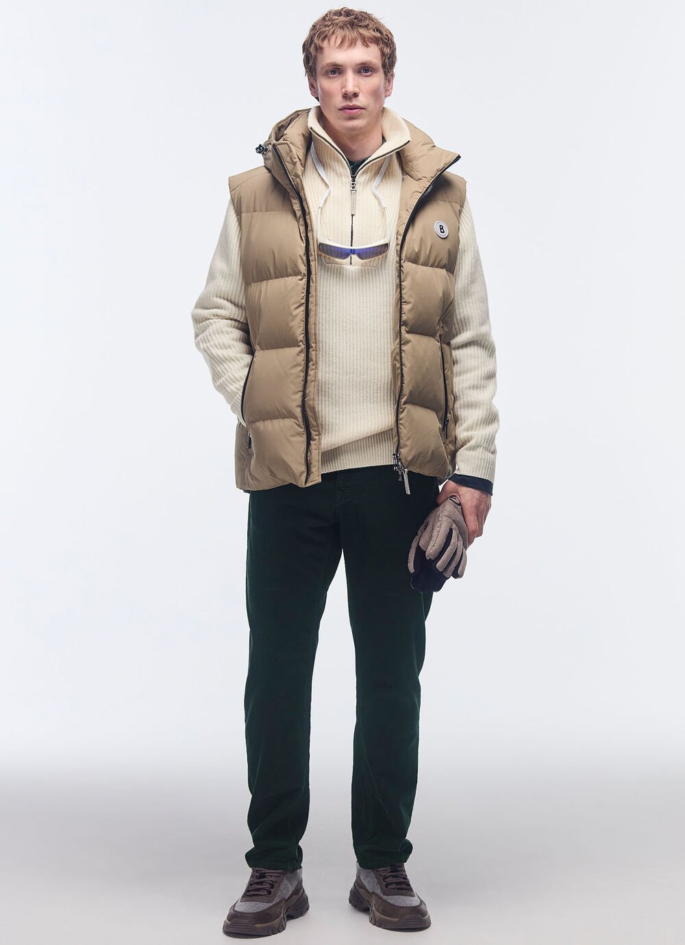 Het model draagt een camelkleurige donzen bodywarmer met een crèmekleurige trui en een zwarte broek.