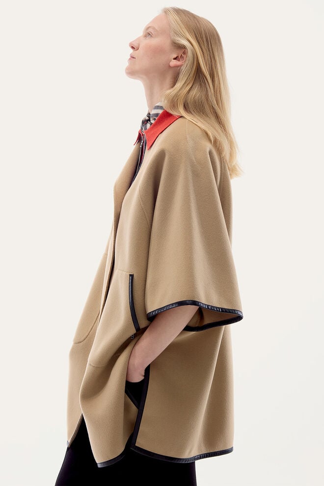 The model is seen from the side wearing a beige cape with black leather trim.