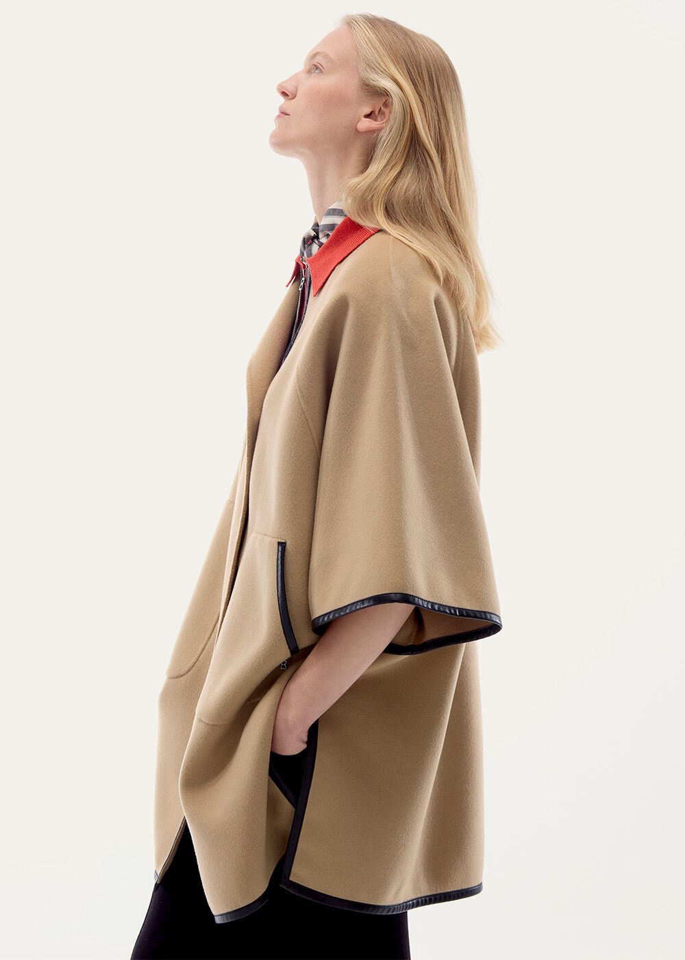 The model is seen from the side wearing a beige cape with black leather trim.