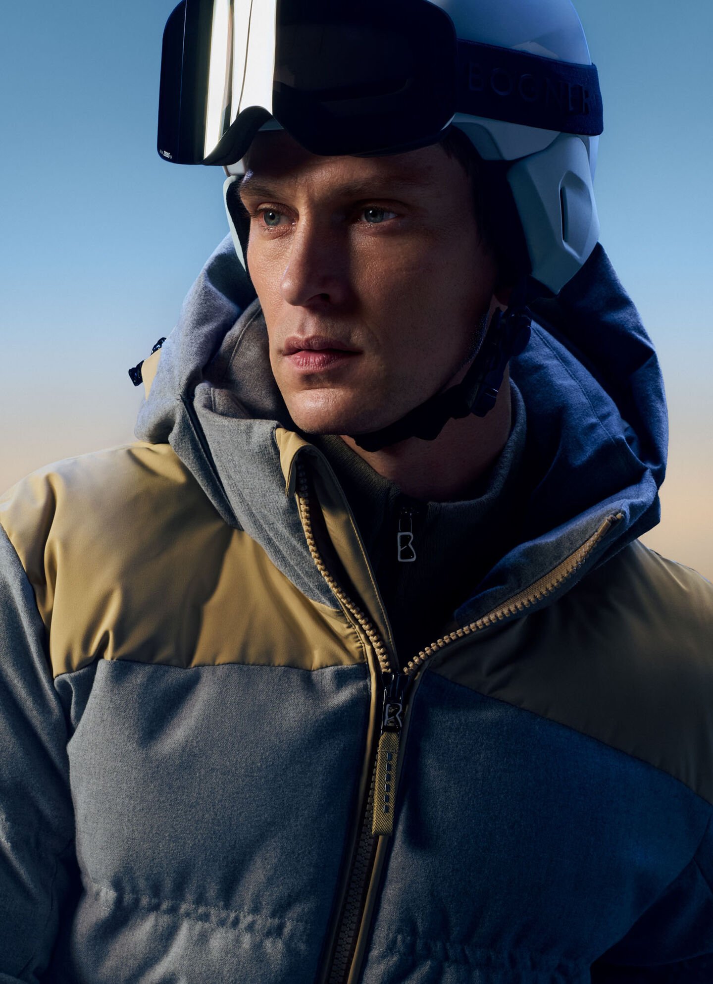 Das Model trägt eine Skidaunenjacke in Grau mit beigefarbenen Einsätzen und einen Skihelm. 