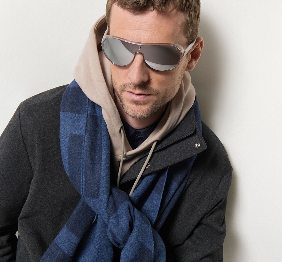 Close-up of the male model. He is wearing polarised sunglasses and a blue striped scarf.