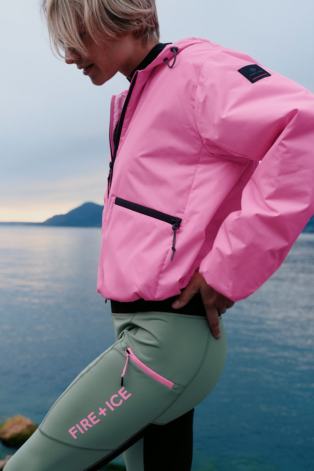 The female model is wearing a pink windbreaker jacket with tights in eucalyptus green. A mountain lake can be seen in the background.