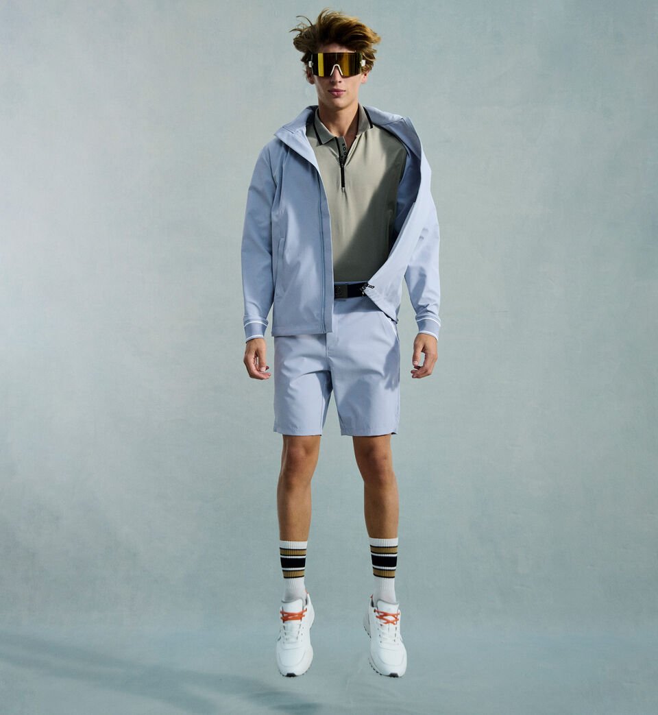 Das Herren Model in dynamischer Pose tr&auml;gt ein eukalyptusfarbenes Polo-Shirt, dazu hellblaue Shorts und eine farblich passende Jacke.