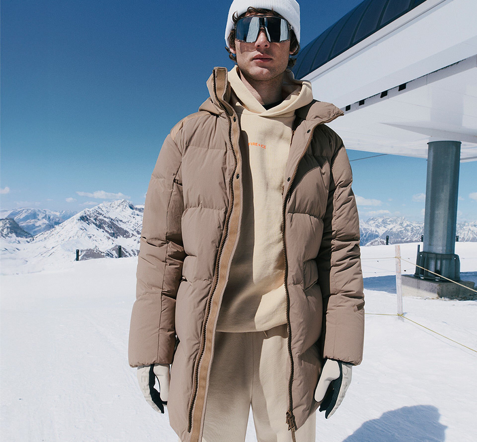  The male model wears a light brown down coat in front of a snow-covered mountain backdrop.
