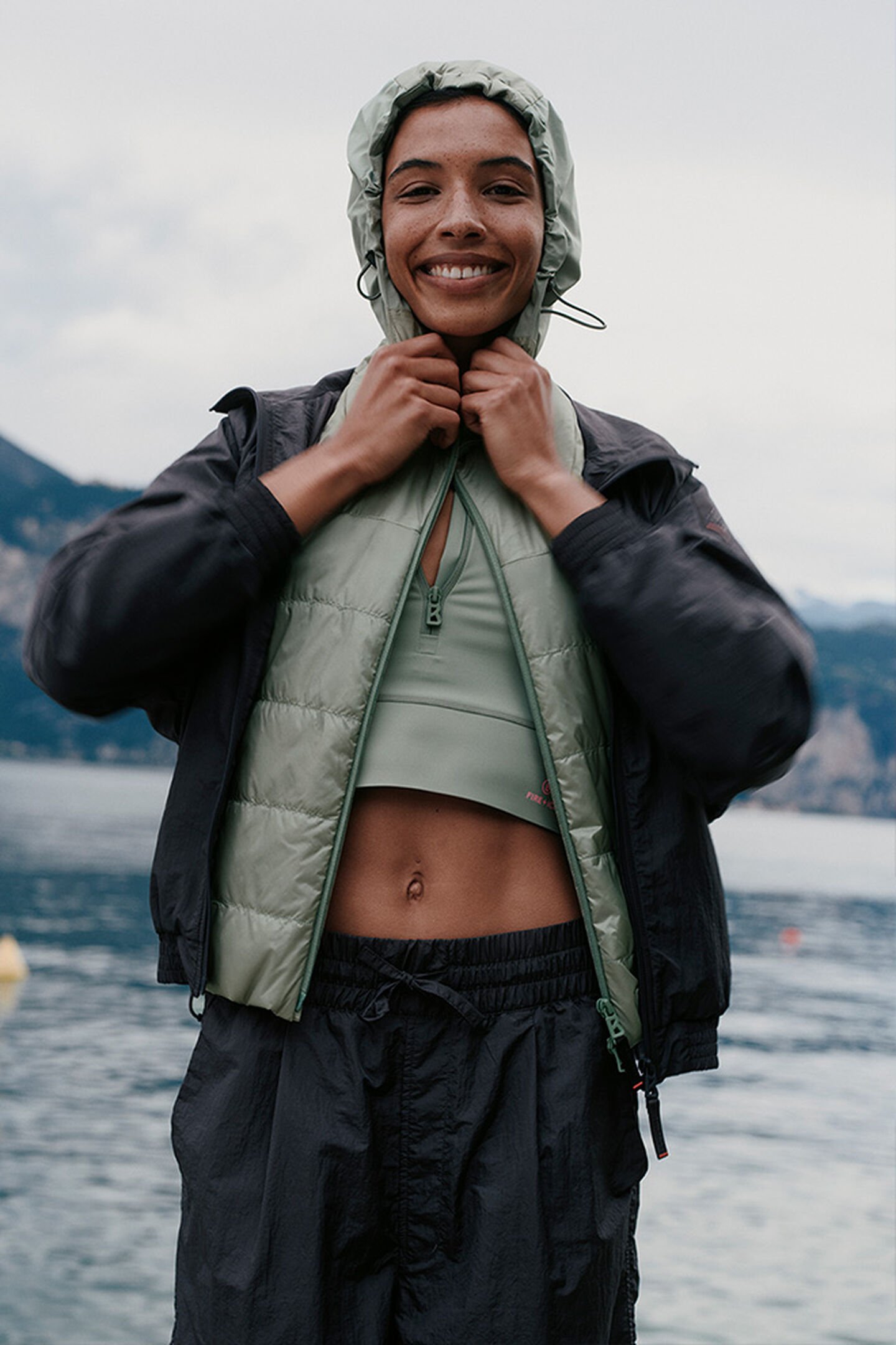 A smiling female model with a green quilted gilet with the hood up. Lake in the background.