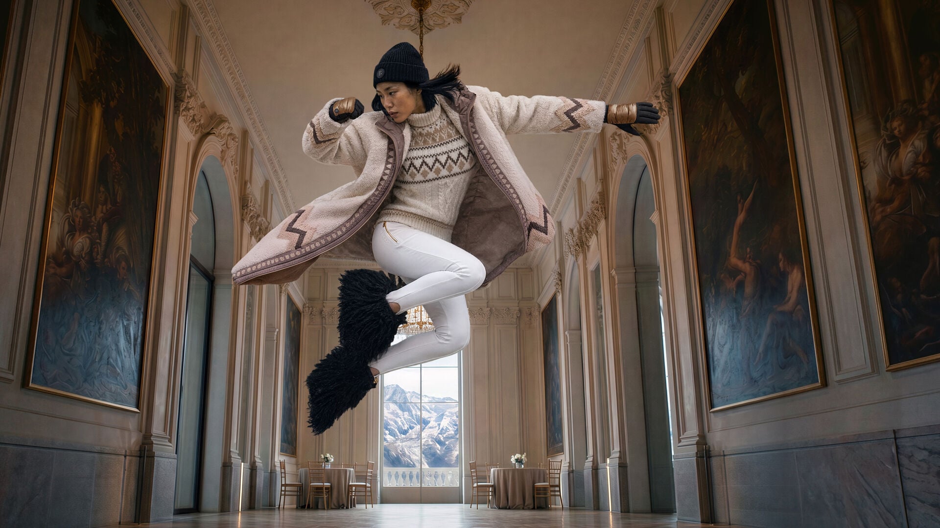 Das Model in dynamischer Pose tr&auml;gt einen gemusterten Mantel &uuml;ber einem Norwegerpullover zu wei&szlig;er Steghose und Fellstiefeln vor alpiner Bergkulisse.