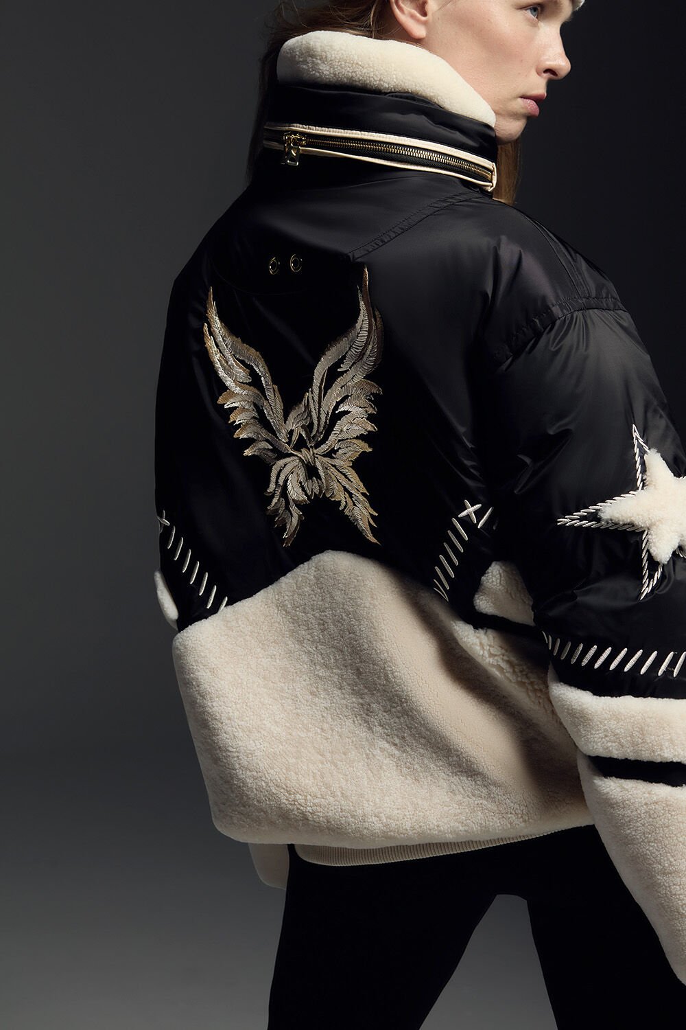 Das Model zeigt die Rückseite ihrer Jacke, die mit Shearling und bestickten Details versehen ist.