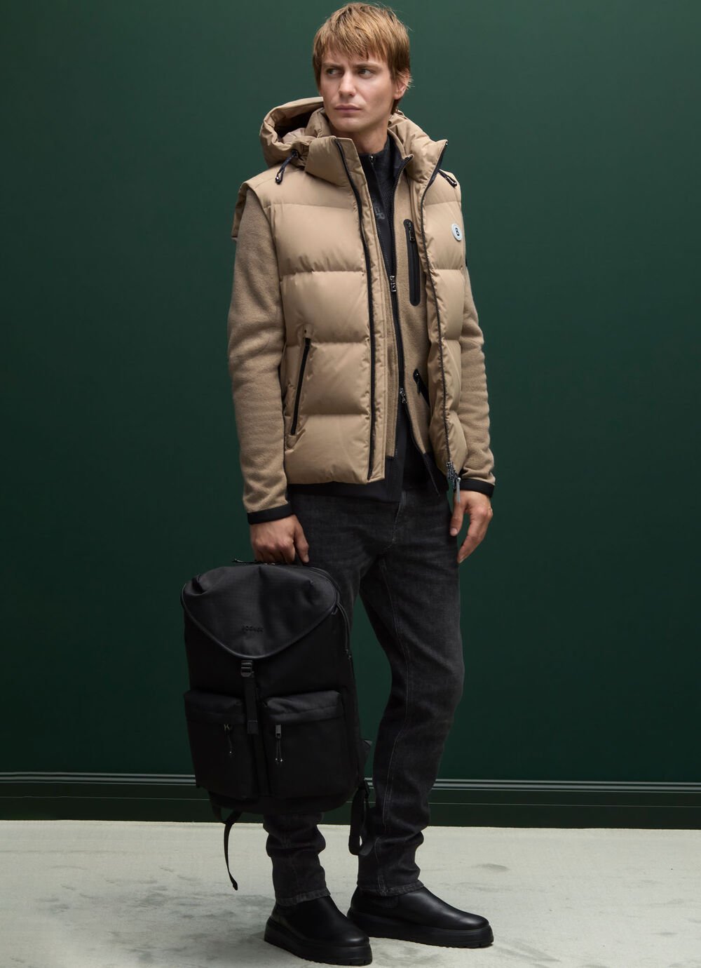 Ce look homme combine un midlayer en laine confortable, une doudoune beige élégante par-dessus, et un sac à dos noir polyvalent.