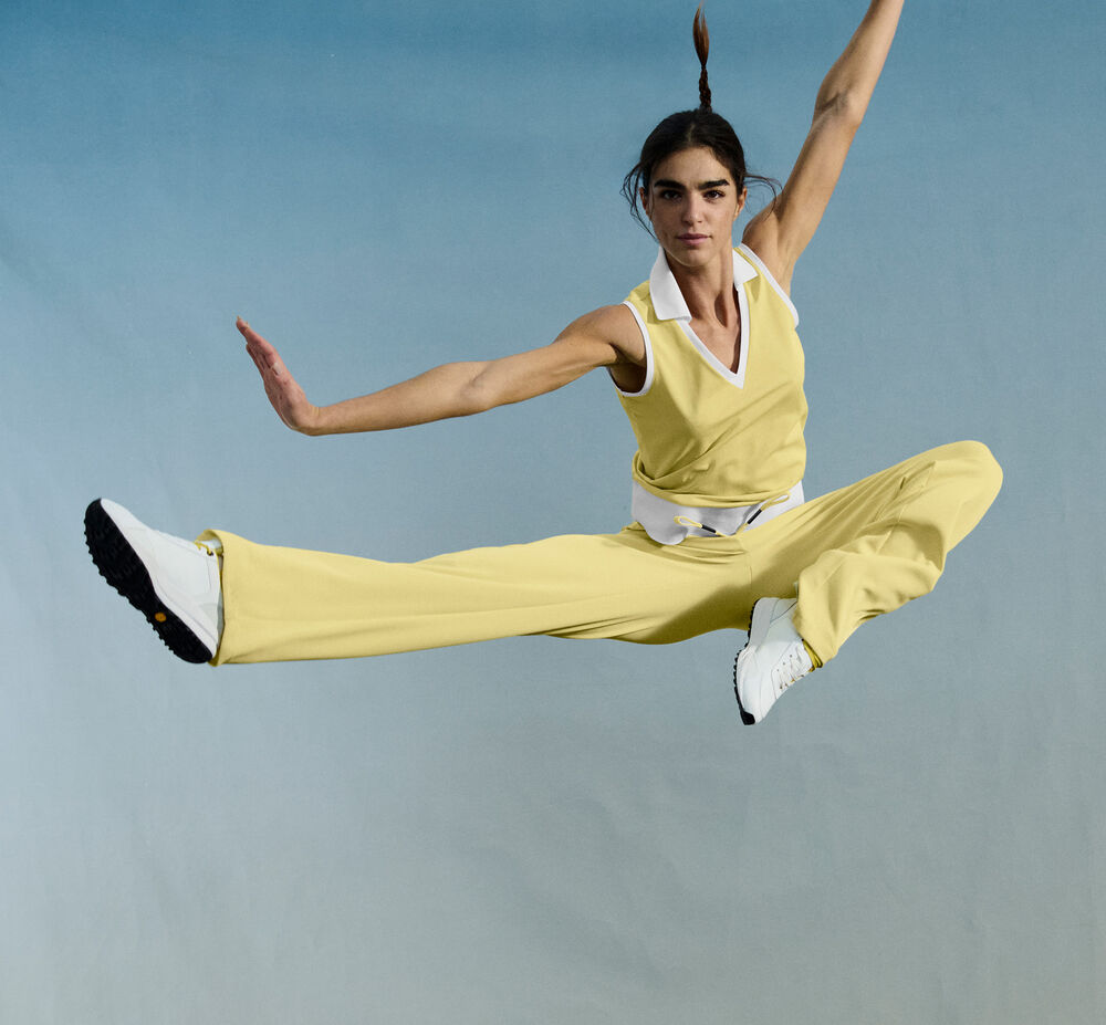 The dark-haired model is seen jumping in the air, wearing a top and long pants in yellow and white.