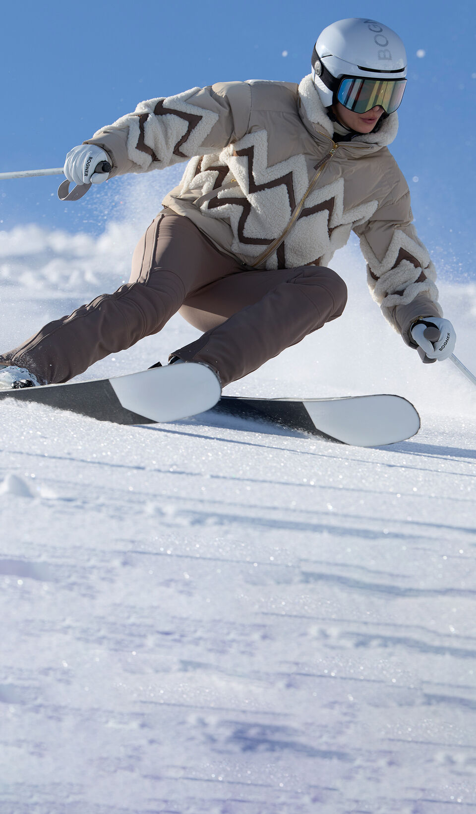 The image shows a model skiing down a freshly groomed slope. She is wearing a ski jacket with a zigzag pattern and brown ski pants.