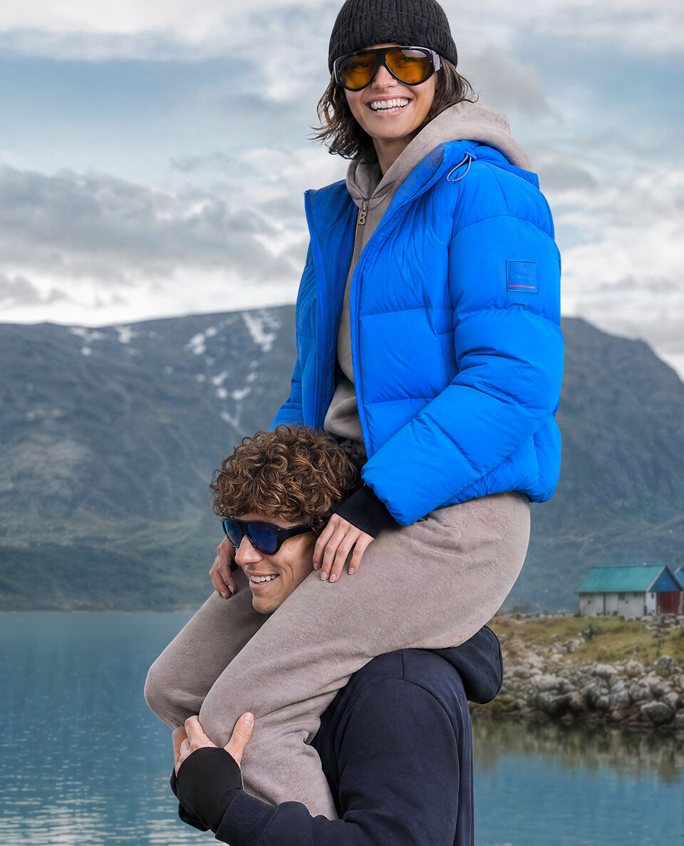 Bergkulisse. Das Herren Model tr&auml;gt das Damen Model auf den Schultern. Sie tr&auml;gt eine royalblaue Steppjacke, eine M&uuml;tze und eine Sonnenbrille.