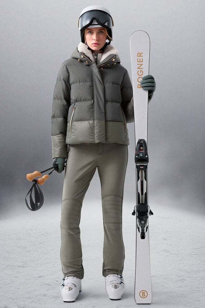 The model is wearing an eucalyptus-colored ski outfit and is holding a ski in her hand.