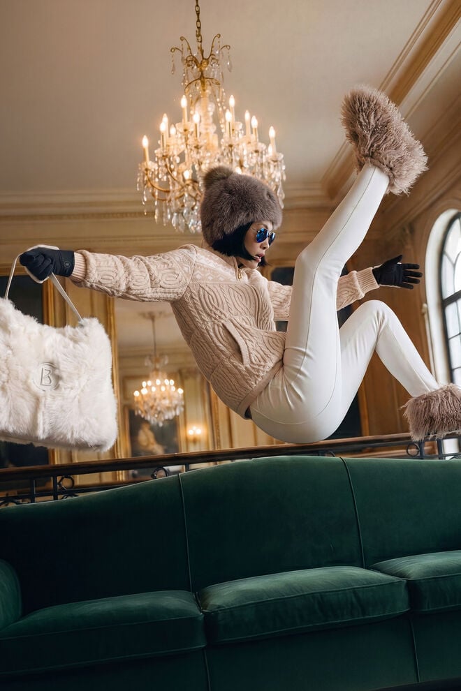 Das Damen Model in dynamischer Pose vor einer Hotelkulisse tr&auml;gt eine gemusterte braune Strickjacke, dazu eine wei&szlig;e Steghose, Fellboots und Fellm&uuml;tze. Sie h&auml;lt eine mit Fell besetzte Tasche in der Hand.