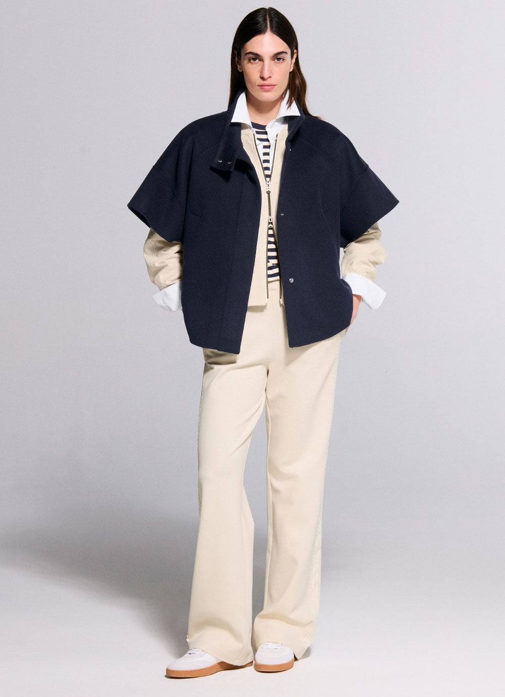 Das Model tr&auml;gt ein cremefarbenes Outfit, darunter ein gestreiftes T-Shirt. Dar&uuml;ber tr&auml;gt sie einen marineblauen Woll-Poncho.