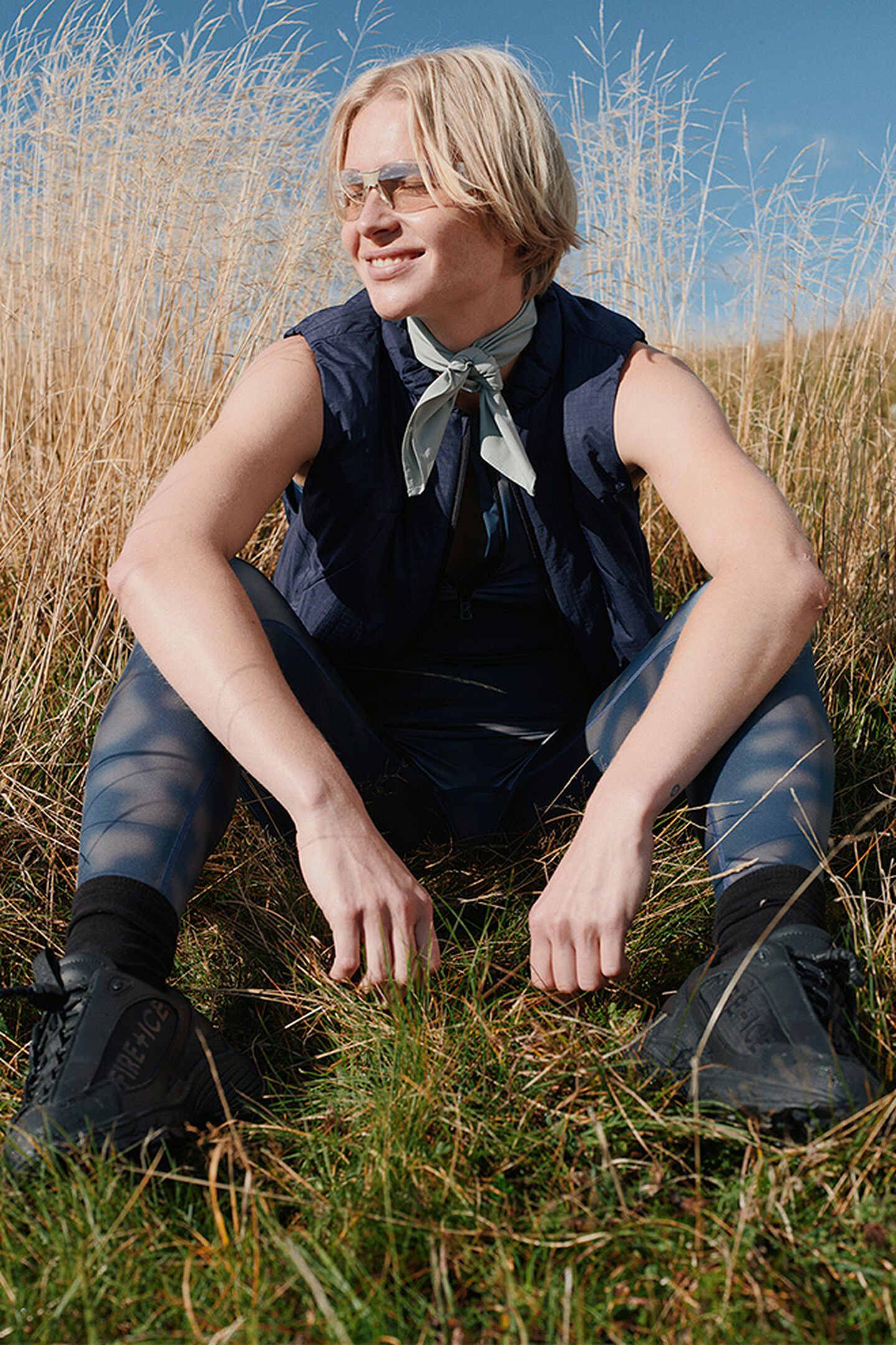 A female model sitting in a meadow landscape. She is wearing a sleeveless gilet, paired with printed tights and FIRE+ICE trainers.