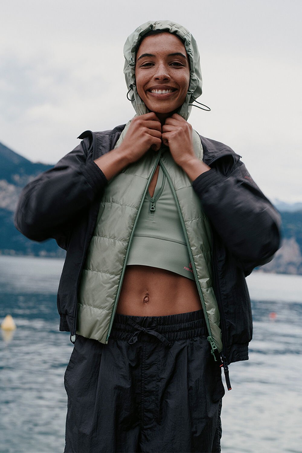 Ein l&auml;chelndes Damen Model mit gesteppter gr&uuml;ner Weste, deren Kapuze sie &uuml;ber den Kopf gezogen hat. See im Hintergrund.