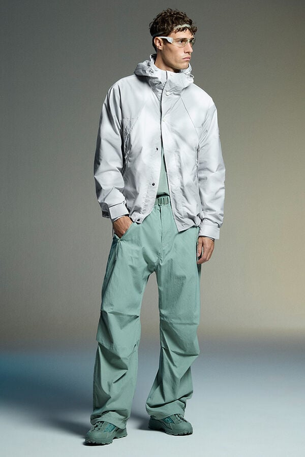 Das Herren Model tr&auml;gt eine wei&szlig;-grau gemusterte Softshell-Jacke, dazu eine lange, leichte Hose in Gr&uuml;n. 