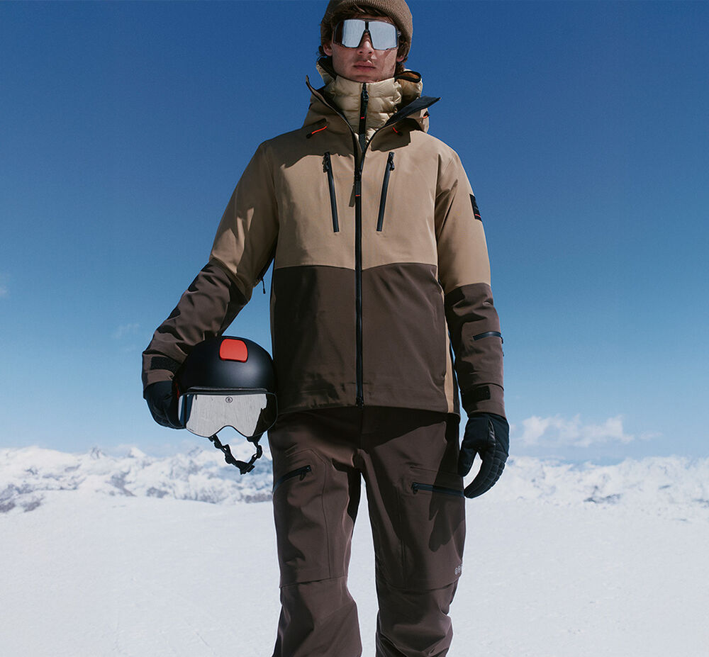 Das Herren Model trägt einen in Brauntönen gehaltenen Ski Look, der mit einer Skibrille, einem Helm und Handschuhen ergänzt wird.